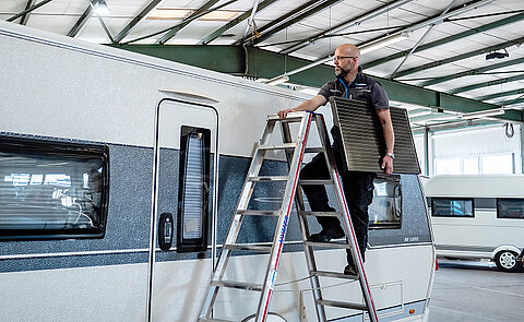 Werkstattmitarbeiter von Caravaning Center Schmidtmeier auf einer Leiter an einem Wohnwagen und einer Solar-Paneele unter dem Arm.
